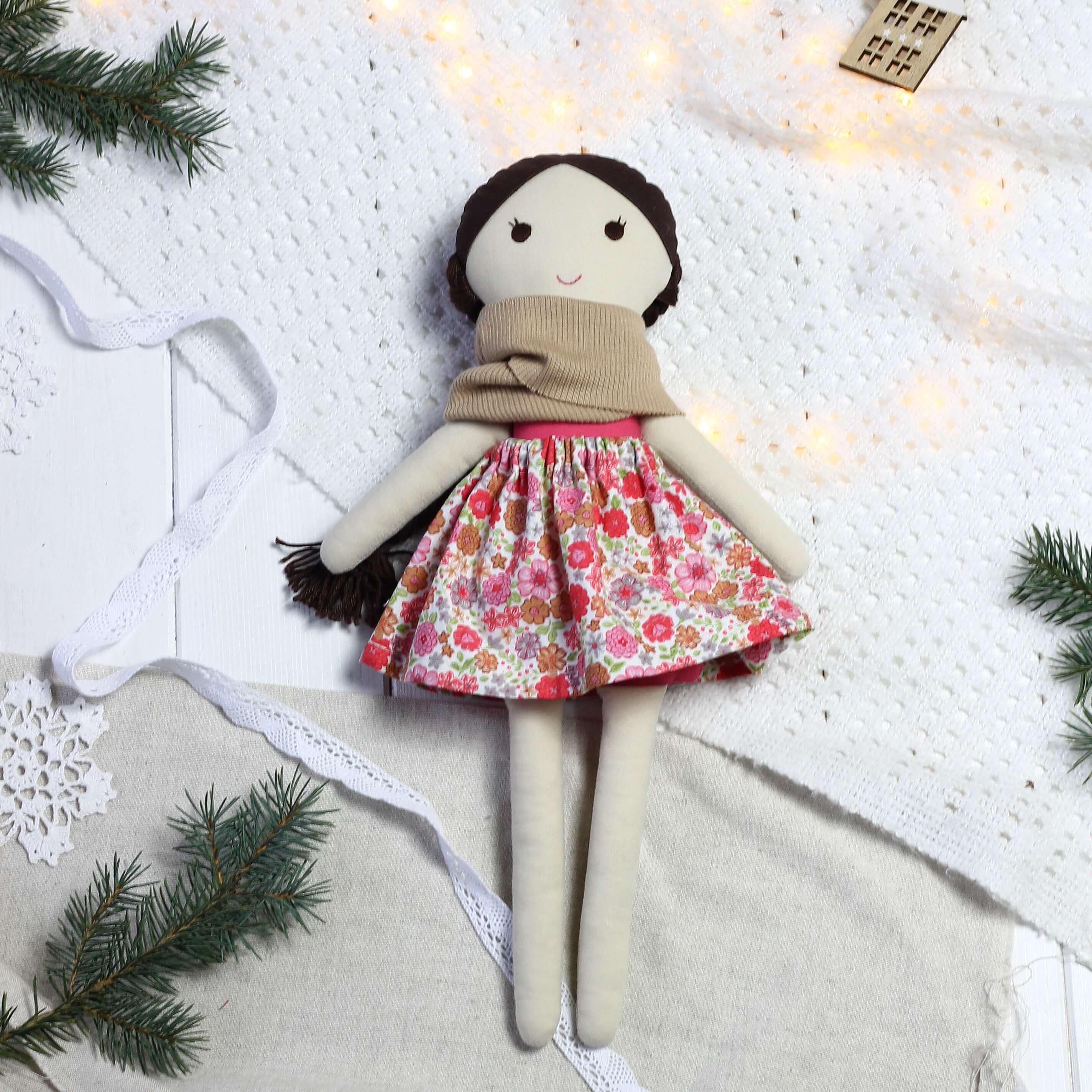 Ręcznie szyta lalka handmade w kwiecistej spódniczce i beżowym kominie, ułożona na jasnym tle